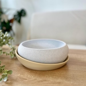 May include: A light gray, ribbed ceramic bowl sits atop a light yellow saucer. The bowl has a rounded shape with vertical grooves. The saucer is a simple, shallow dish. The items are on a wooden surface.