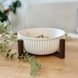 May include: A white, ribbed ceramic bowl on a dark brown wooden stand. The bowl has a rounded shape and is filled with small, brown objects. The stand has a simple, geometric design. The bowl and stand are placed on a light brown wooden surface.