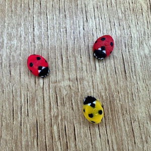 May include: Three ladybug-shaped ornaments, one red with black spots, one red with black spots, and one yellow with black spots, on a woodgrain background.