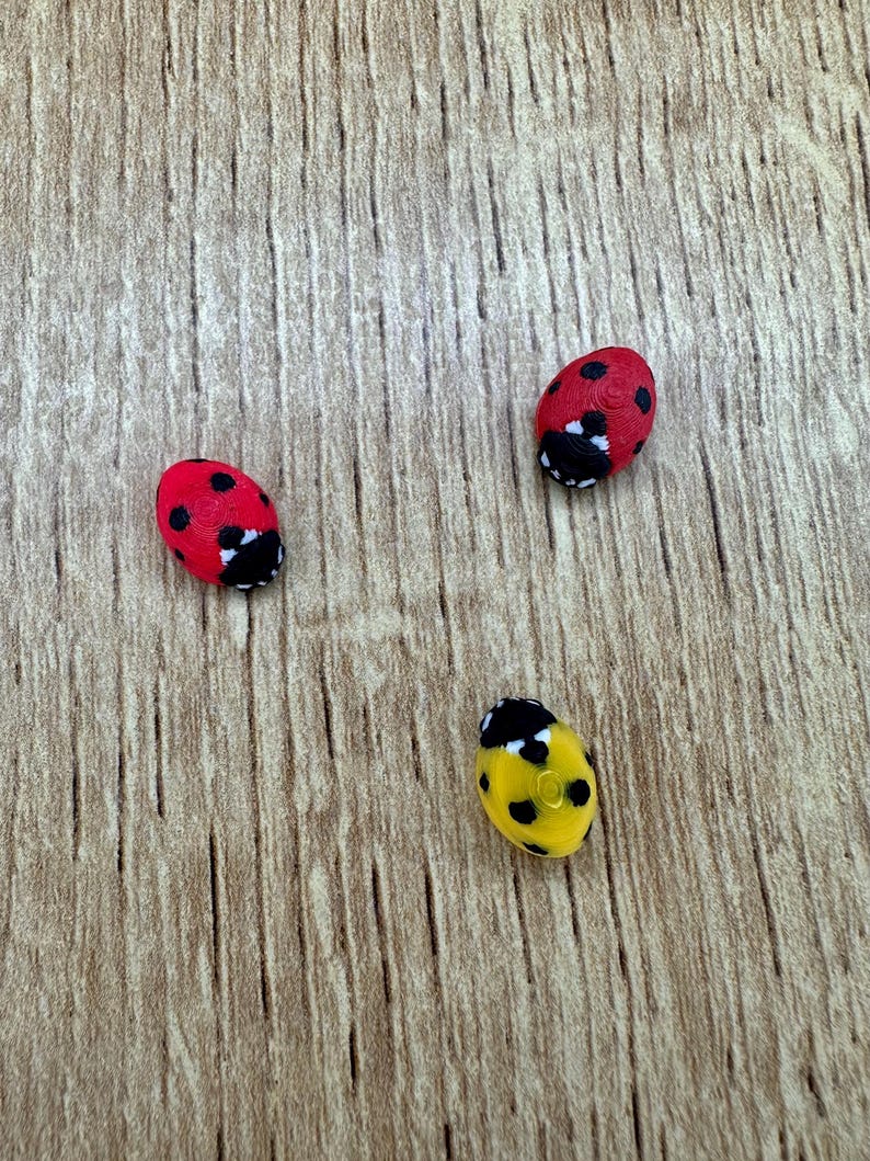 May include: Three ladybug-shaped ornaments, one red with black spots, one red with black spots, and one yellow with black spots, on a woodgrain background.