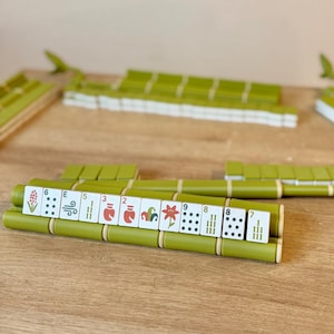 May include: A set of Mahjong tiles arranged on green bamboo-style racks. The white tiles feature various colorful illustrations, including flowers and numbers, alongside traditional dot and bamboo patterns. The set is organized in rows on a wooden surface.