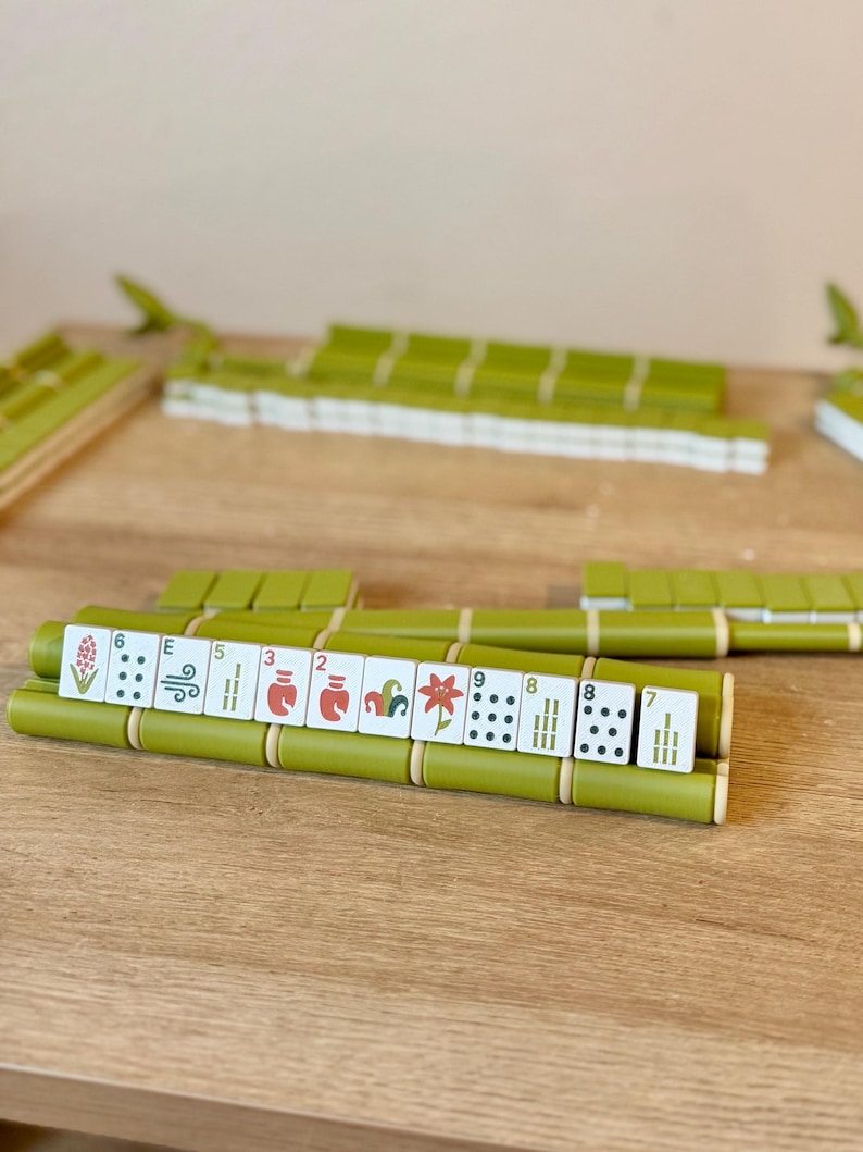 May include: A set of Mahjong tiles arranged on green bamboo-style racks. The white tiles feature various colorful illustrations, including flowers and numbers, alongside traditional dot and bamboo patterns. The set is organized in rows on a wooden surface.