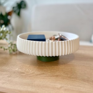 May include: A cream-colored, ribbed decorative tray with a green base holds a small blue box and keys. The tray sits on a light brown wooden surface. The tray's design features vertical ridges around its perimeter.