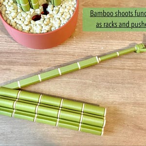 May include: A green, segmented bamboo-shaped tool with white rings, designed to function as a rack and pusher. The object features a long, slender arm extending from a base of three parallel, connected cylindrical segments with a leaf-like tip.