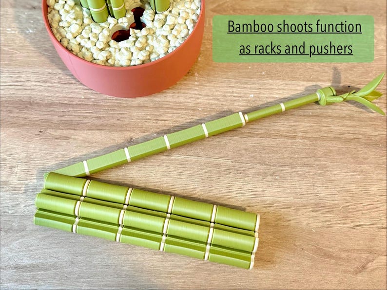 May include: A green, segmented bamboo-shaped tool with white rings, designed to function as a rack and pusher. The object features a long, slender arm extending from a base of three parallel, connected cylindrical segments with a leaf-like tip.