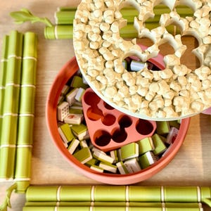 May include: A round terracotta-colored container filled with small green and white rectangular tiles and a pink insert, topped with a textured beige circular lid. The set is surrounded by four green bamboo-shaped rods with white accents.