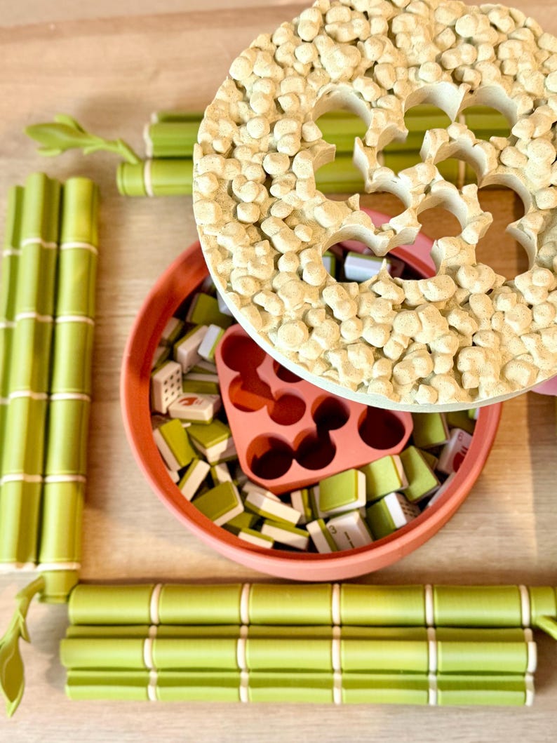 May include: A round terracotta-colored container filled with small green and white rectangular tiles and a pink insert, topped with a textured beige circular lid. The set is surrounded by four green bamboo-shaped rods with white accents.