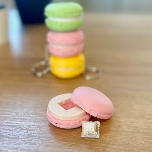 May include: A pink macaron shaped keycap holder with a white keycap inside. The holder is open and the keycap is visible. There are also three stacked macarons in the background, one green, one pink, and one yellow.