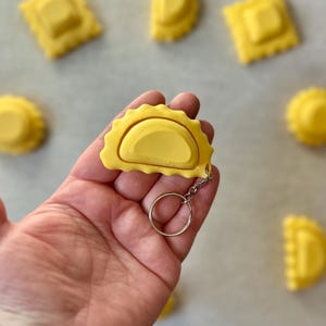 May include: A yellow ravioli keychain with a silver key ring. The keychain is shaped like a half-moon ravioli with a raised center.
