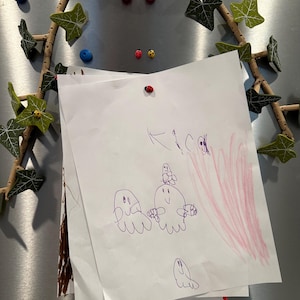 May include: A white sheet of paper with a purple drawing of three ghosts and a pink scribble in the background. The paper is attached to a silver refrigerator with a red ladybug magnet.