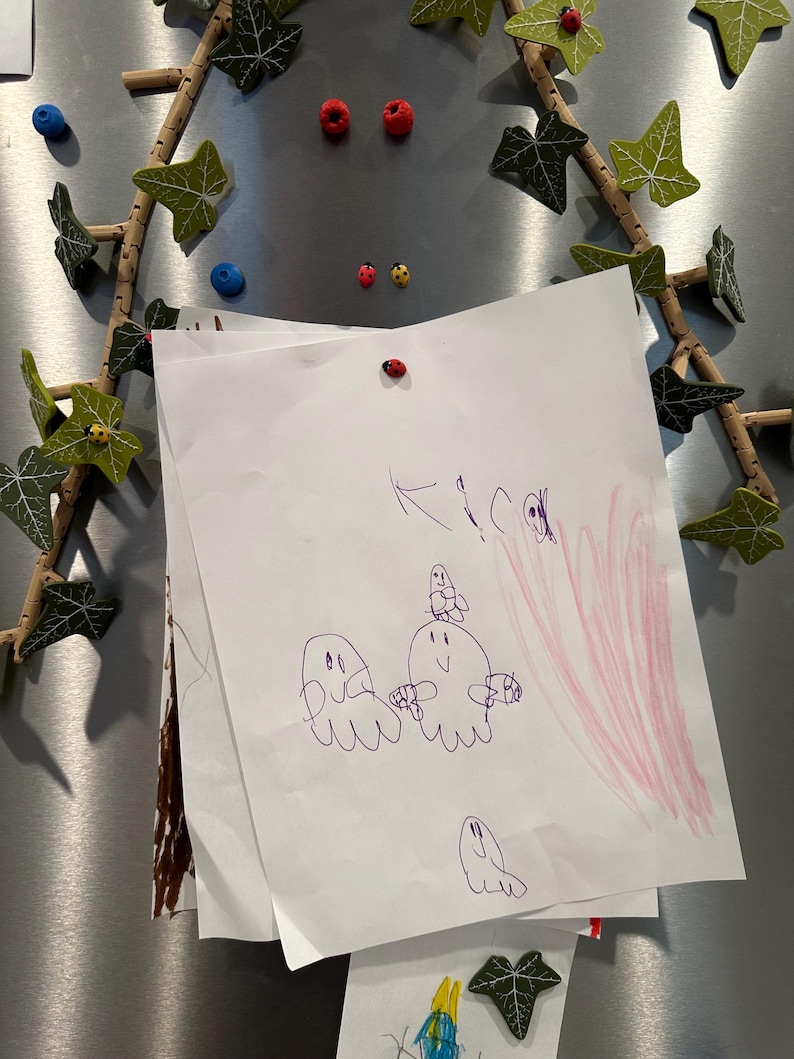 May include: A white sheet of paper with a purple drawing of three ghosts and a pink scribble in the background. The paper is attached to a silver refrigerator with a red ladybug magnet.
