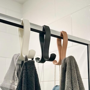 May include: Three shower hooks in white, black, and tan hang from a black bar. Each hook holds a towel. The towels are gray and dark gray. A blue bath sponge hangs from one of the hooks. The background is white tile.