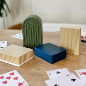 May include: A collection of playing card holders in various colors and shapes. There is a dark green arch-shaped holder, a beige rectangular holder, a navy blue rectangular holder, and a beige square holder. Playing cards are scattered around the holders on a wooden table.