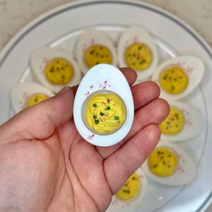 May include: A plate of deviled eggs with a single egg held in a hand. The egg is topped with a yellow yolk mixture and green chives.