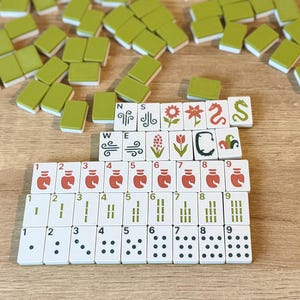 May include: A set of Mahjong tiles featuring white faces with various printed designs including numbers, dots, bamboo, dragons, and floral motifs, alongside plain olive green tiles scattered around the organized rows of game pieces.