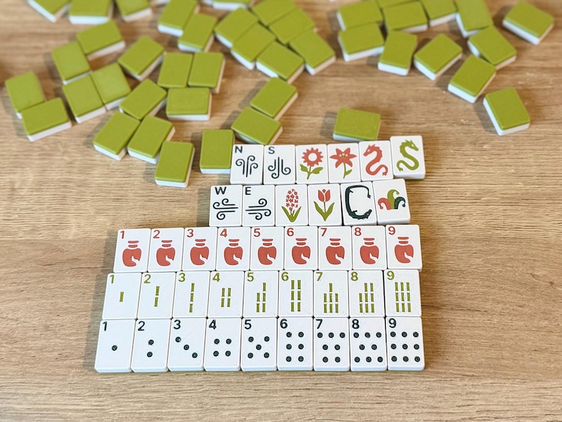 May include: A set of Mahjong tiles featuring white faces with various printed designs including numbers, dots, bamboo, dragons, and floral motifs, alongside plain olive green tiles scattered around the organized rows of game pieces.