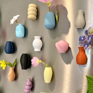 May include: A collection of 12 small, colorful, 3D printed vases with various shapes and textures. Some vases are filled with small flowers. The vases are attached to a silver refrigerator door.