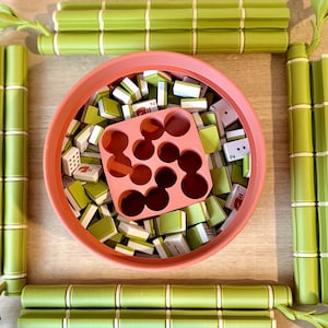 May include: A round coral-colored bowl filled with small rectangular game tiles featuring green and white patterns, accompanied by a square coral-colored insert with circular cutouts. Four green bamboo-shaped game board segments surround the bowl.