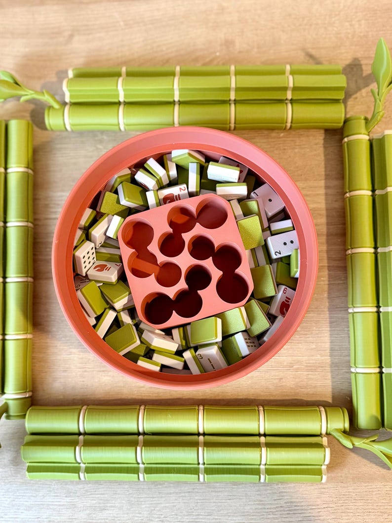 May include: A round coral-colored bowl filled with small rectangular game tiles featuring green and white patterns, accompanied by a square coral-colored insert with circular cutouts. Four green bamboo-shaped game board segments surround the bowl.
