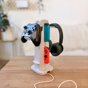 May include: A light gray gaming console stand holds a black controller, teal and red Joy-Cons, and dark green headphones. The stand is on a wooden surface, with a white cable extending from the base.