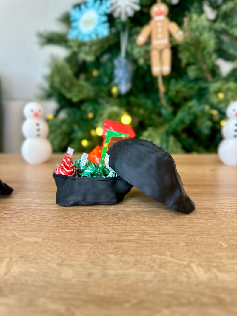 Op de afbeelding: Een zwarte stoffen doos met een deksel, gevuld met rode, groene en witte chocolade snoepjes. De doos staat op een houten oppervlak.