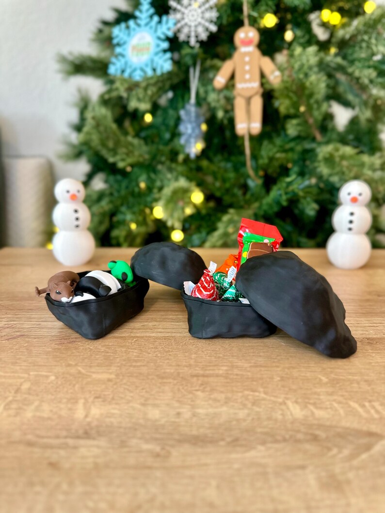 Op de afbeelding: Twee zwarte, rotsvormige dozen met open deksels. E&eacute;n doos bevat een klein knuffeldier en een gestreept speelgoed. De andere doos bevat kleurrijke verpakte snoepjes.