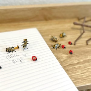 May include: A notebook with the handwritten text "Office Bugs" and several miniature insect decorations. The insects include bees, ladybugs, and a stick insect, arranged on a wooden surface.