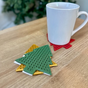 May include: Three Christmas tree-shaped coasters. The top coaster is green with a woven pattern. The middle coaster is white. The bottom coaster is gold. The coasters are on a wooden surface.
