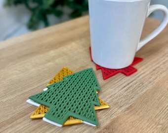 May include: Three Christmas tree-shaped coasters. The top coaster is green with a woven pattern. The middle coaster is white. The bottom coaster is gold. The coasters are on a wooden surface.