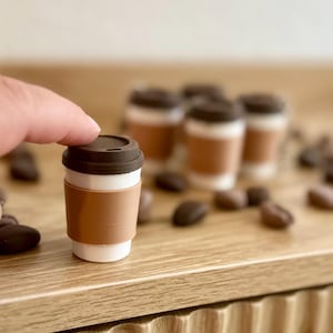May include: Miniature coffee cups with brown lids and sleeves, arranged on a wooden surface with scattered coffee beans. The cups have a white base and a brown sleeve. A finger is touching the lid of one cup.