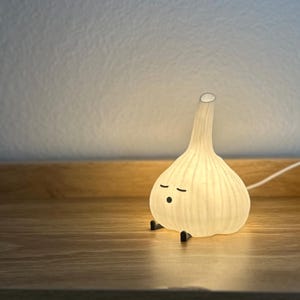 May include: A whimsical, illuminated garlic bulb-shaped lamp with a smiling face and small black feet. The lamp emits a soft, warm glow, creating a cozy ambiance. The lamp is sitting on a wooden surface.
