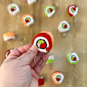 May include: A hand holds a red, orange, and green sushi keychain. The keychain is shaped like a sushi roll and has a white rice filling with a green avocado and a red fish topping. The keychain is attached to a silver key ring.