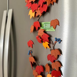 May include: A decorative vine with colorful fall leaves in shades of red, orange, and brown, attached to a refrigerator. The leaves are arranged along a brown stem, with small blue berry clusters. A small green card with text is also attached.