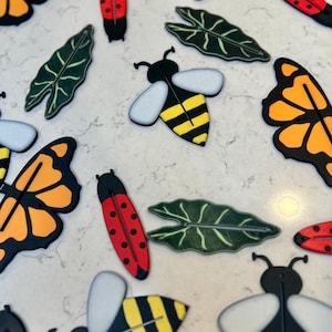 May include: A collection of colorful wooden cutouts of butterflies, bees, ladybugs, and leaves. The cutouts are painted in bright colors and have a whimsical, nature-inspired design.