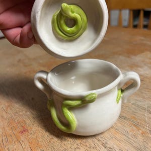 May include: A white ceramic cup with two handles and a green snake wrapped around the side. The lid, held above, features a coiled green snake. The cup is on a wooden surface.