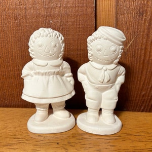 May include: Two unpainted ceramic figurines of a girl and a boy. The girl wears a dress and has curly hair. The boy wears a sailor hat and has his hands in his pockets. Both have smiling faces and are on oval bases.