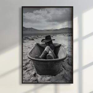 May include: A black and white photograph of a person in a cowboy hat sitting in a bathtub in a desert landscape.