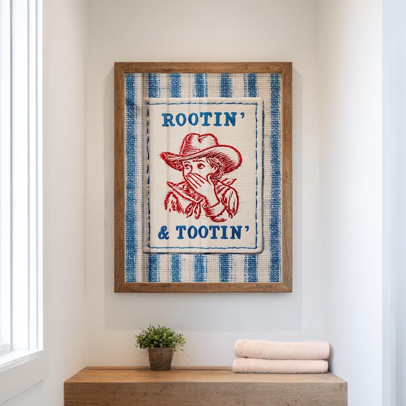 Framed “Rootin’ & Tootin’” red and blue cowboy wall art hanging above a toilet in a bright, neutral-toned bathroom with soft natural light and cozy, rustic decor.