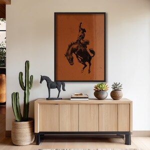 May include: A framed print featuring a black and white illustration of a cowboy riding a bucking bronco. The artwork is set against a rust-colored background. A wooden cabinet with a black metal base holds decorative items, including a cactus and a horse statue.
