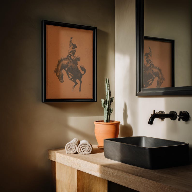 May include: A framed print of a cowboy riding a bucking bronco hangs above a bathroom sink. The print is in a black frame and features a sepia-toned image. A cactus in a terracotta pot and rolled towels sit on the wooden counter.