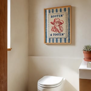 Framed “Rootin’ & Tootin’” red and blue cowboy wall art hanging above a toilet in a bright, neutral-toned bathroom with soft natural light and cozy, rustic decor.