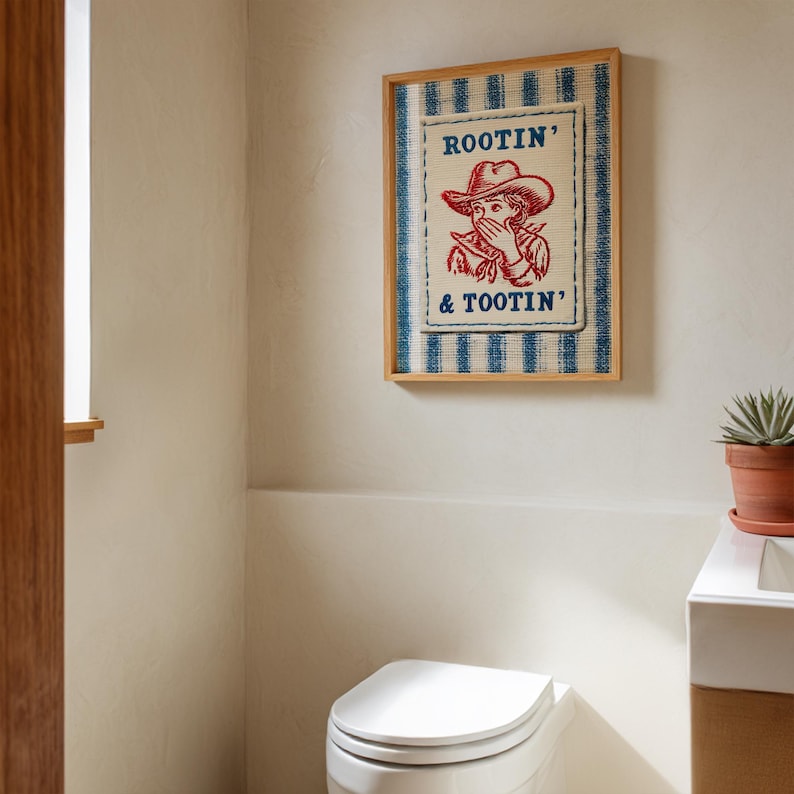 Framed “Rootin’ & Tootin’” red and blue cowboy wall art hanging above a toilet in a bright, neutral-toned bathroom with soft natural light and cozy, rustic decor.