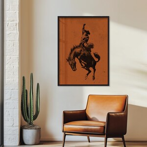May include: A framed print featuring a black and white illustration of a cowboy riding a bucking bronco. The artwork is set against a textured orange background and framed in black. The image evokes a sense of the American West.