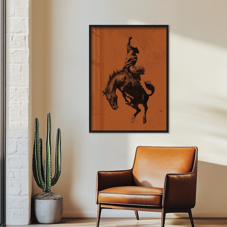 May include: A framed print featuring a black and white illustration of a cowboy riding a bucking bronco. The artwork is set against a textured orange background and framed in black. The image evokes a sense of the American West.