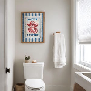 Framed “Rootin’ & Tootin’” red and blue cowboy wall art hanging above a toilet in a bright, neutral-toned bathroom with soft natural light and cozy, rustic decor.