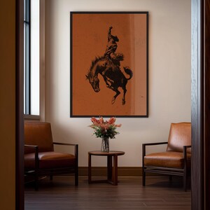 May include: A framed art print featuring a black and brown silhouette of a rodeo cowboy riding a bucking bronco against a burnt orange background. The artwork is displayed in a room with leather chairs, a small table, and a vase of flowers.
