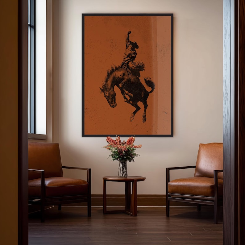 May include: A framed art print featuring a black and brown silhouette of a rodeo cowboy riding a bucking bronco against a burnt orange background. The artwork is displayed in a room with leather chairs, a small table, and a vase of flowers.
