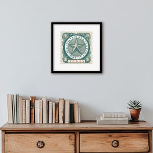K&ouml;nnte beinhalten: Gerahmter Vintage-Kunstdruck mit Texas-Briefmarke. Die Briefmarke zeigt ein zentrales Sterndesign in T&uuml;rkis, umgeben von dem Text "UNITED STATES POSTAL SERVICE" und "TEXAS". Das Kunstwerk ist in einem schwarzen Rahmen gerahmt und auf einer Holzoberfl&auml;che ausgestellt.