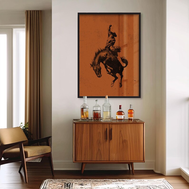 May include: A framed print of a cowboy riding a bucking bronco, in a black frame. The artwork is in shades of brown and black, mounted on an orange background. Below the artwork is a wooden cabinet with several bottles of liquor.