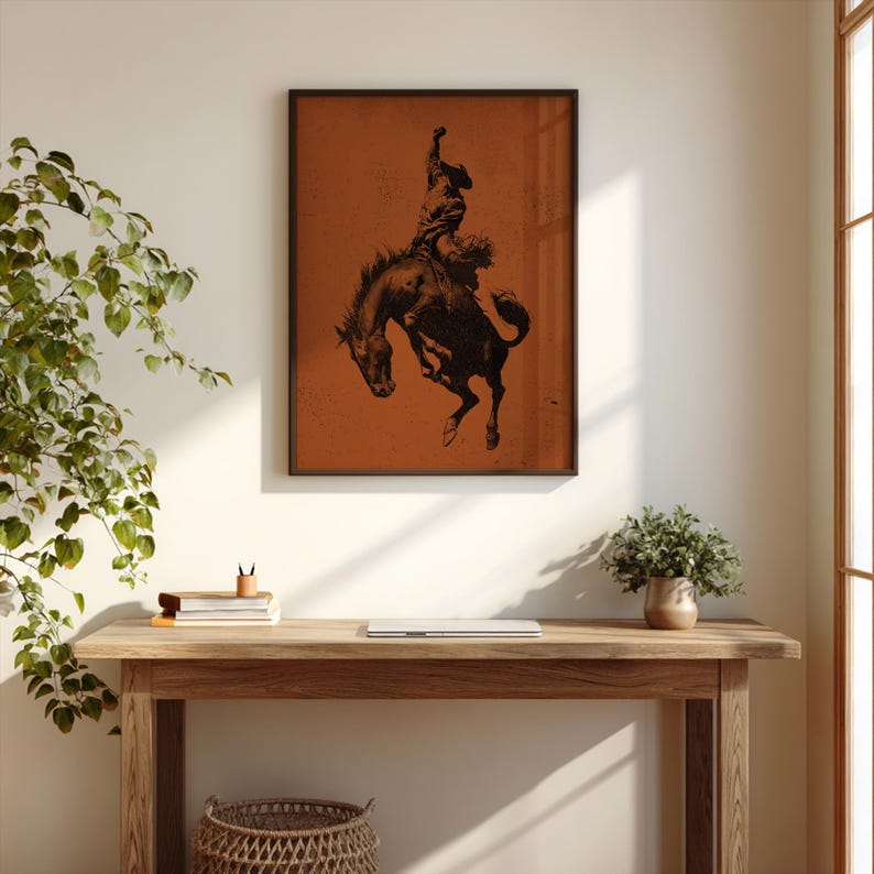 May include: A framed art print featuring a black silhouette of a rodeo cowboy riding a bucking bronco. The artwork has a vintage aesthetic, with a distressed orange background and a dark brown frame. The print is displayed on a wall above a wooden desk.